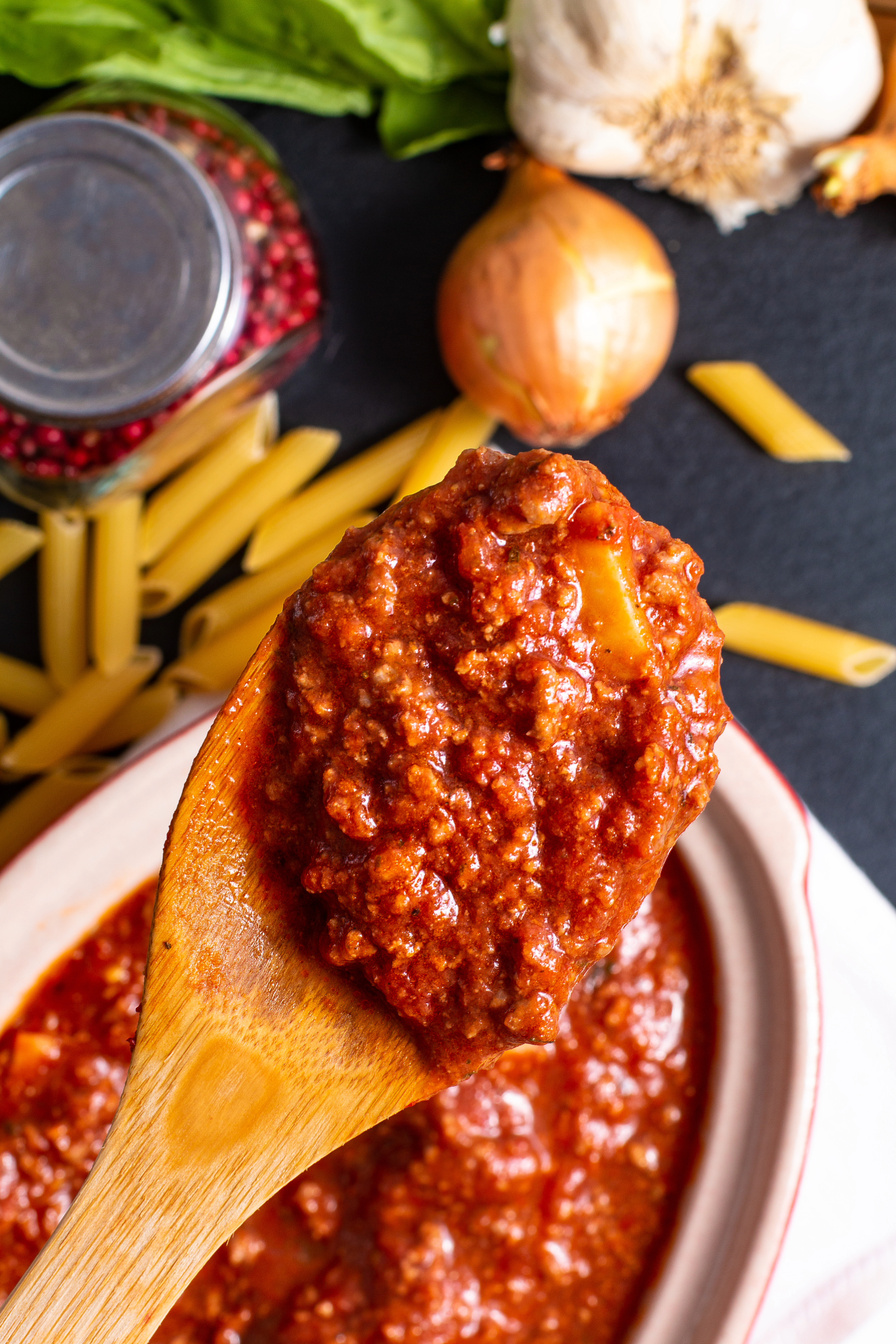 Wooden spoon with tomato sauce over a bowl, surrounded by pasta, garlic, and onions on a dark surface.