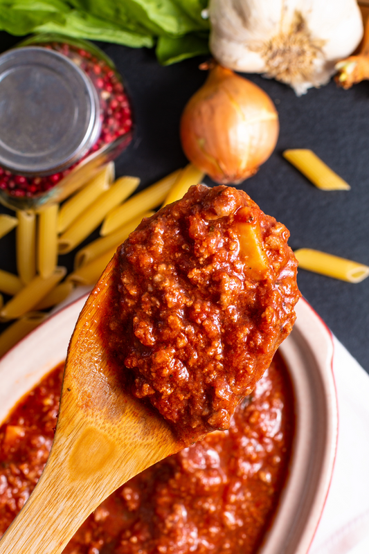 Wooden spoon with tomato sauce over a bowl, surrounded by pasta, garlic, and onions on a dark surface.