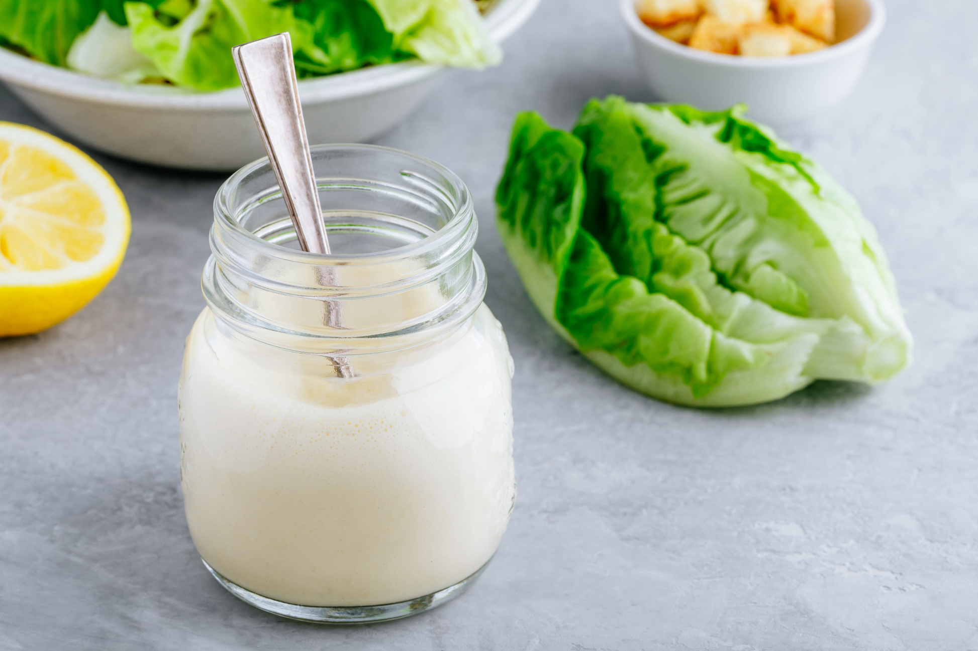 Jar of creamy dressing with a spoon, surrounded by lettuce and lemon on a gray surface