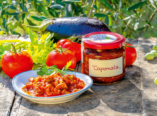 Jar of Caponata with a bowl of pasta and vegetables on a rustic surface with greenery in the background