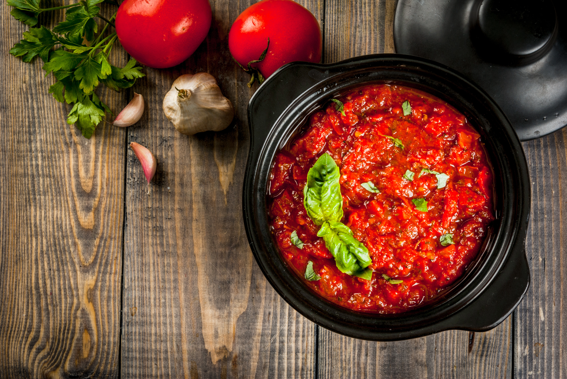 Red sauce in a black pot on a wooden surface with tomatoes and basil.