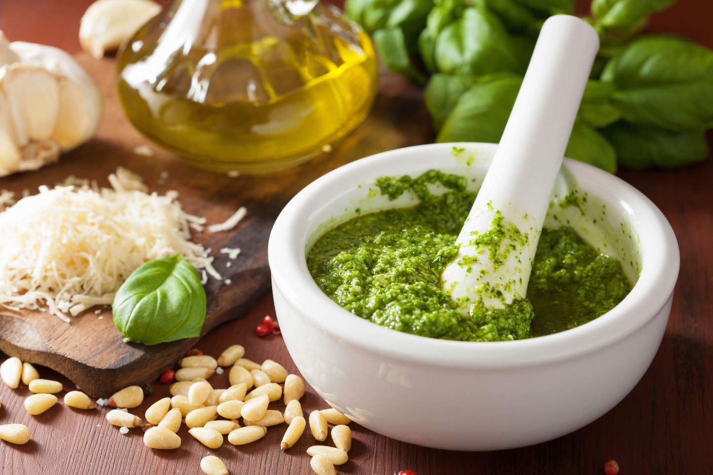 Bowl of green pesto with a white spatula, surrounded by basil, pine nuts, and Parmesan cheese on a wooden surface.